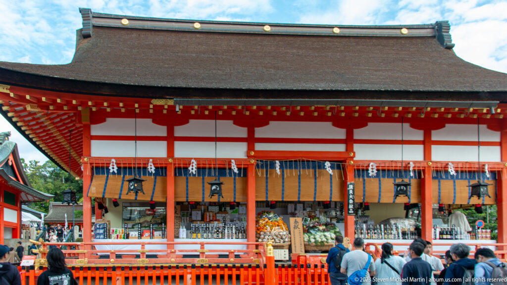 Wide frontal view of a vermilion shrine hall with hanging ropes, lanterns, offerings, and visitors at the railing.