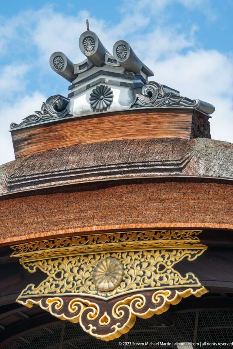 Close-up of an imperial chrysanthemum crest and gold scrollwork beneath a tiled roof ridge.