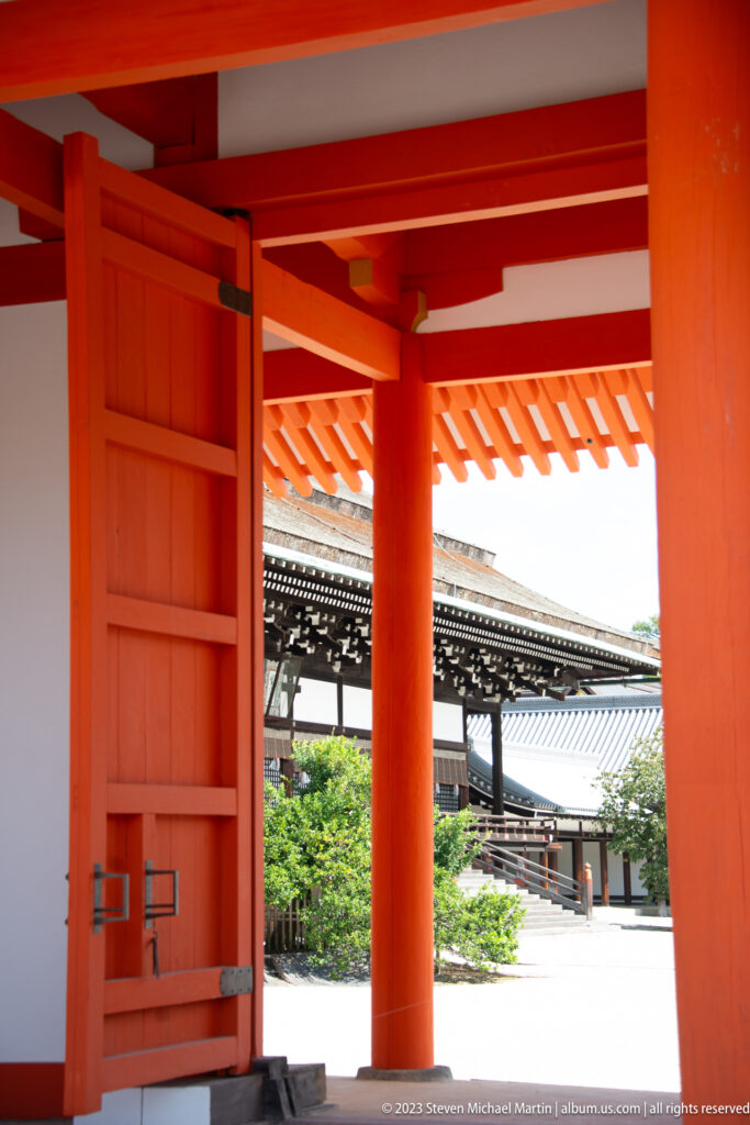 Open vermilion palace gate framing a courtyard, garden, and dark-roofed building beyond.