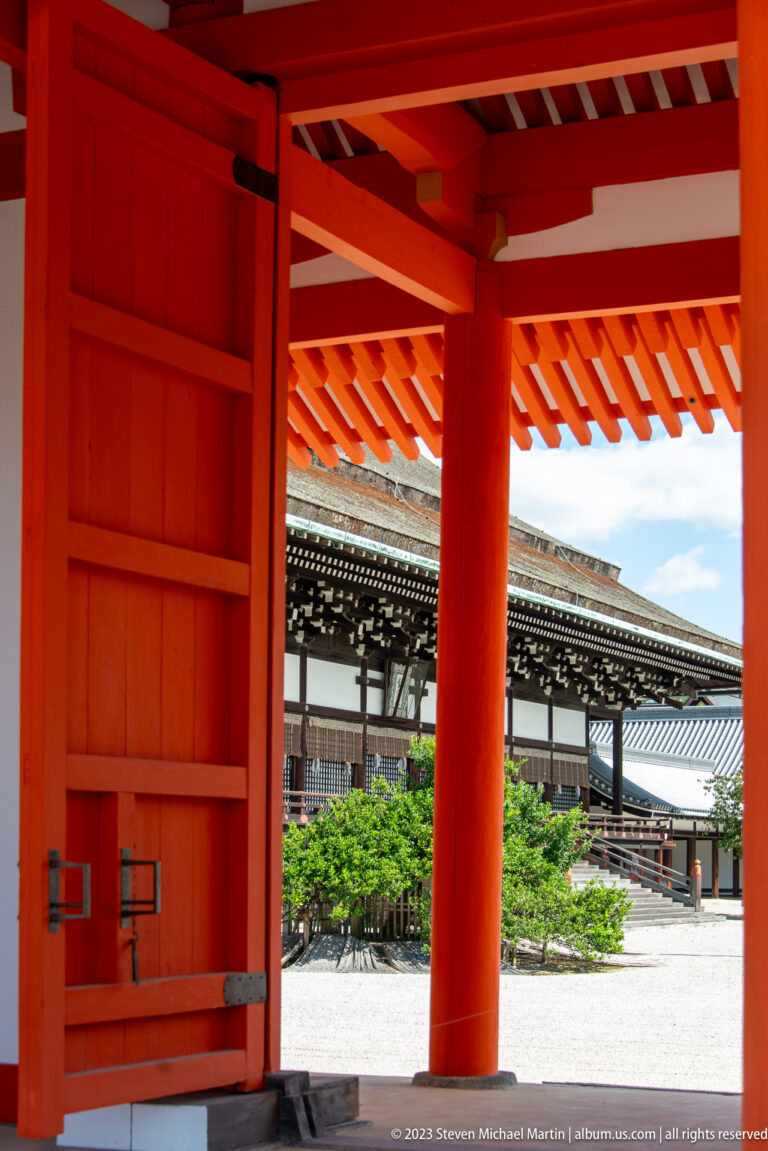Open vermilion gate with visible hardware, garden planting, and roofed palace structure beyond.