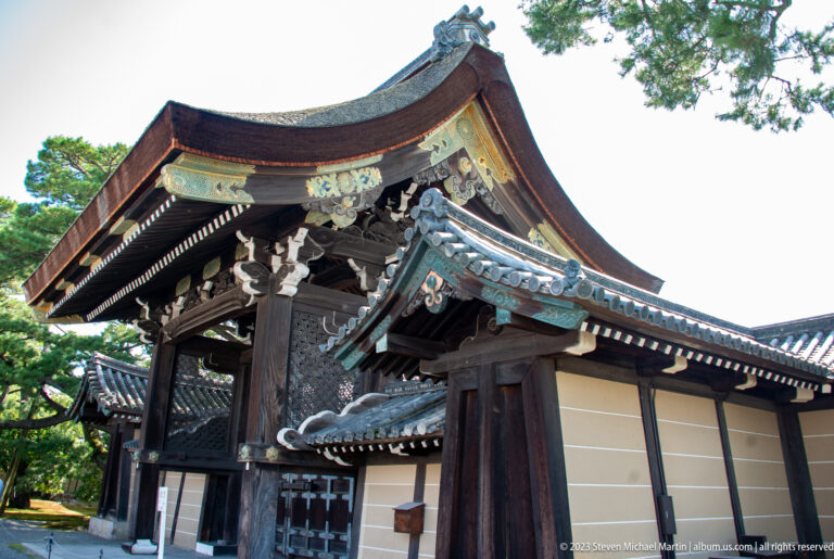 Oblique view of an ornate dark wooden palace gate with layered roofs and trees.