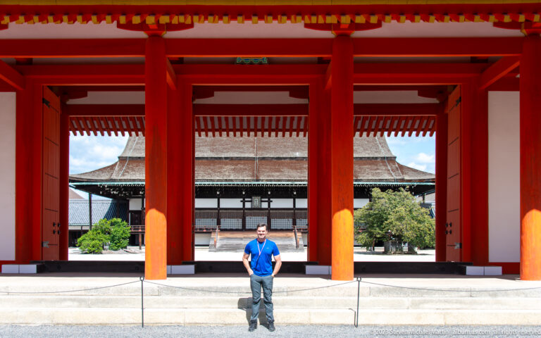 Man standing beneath a large vermilion gate with a palace hall centered behind him.