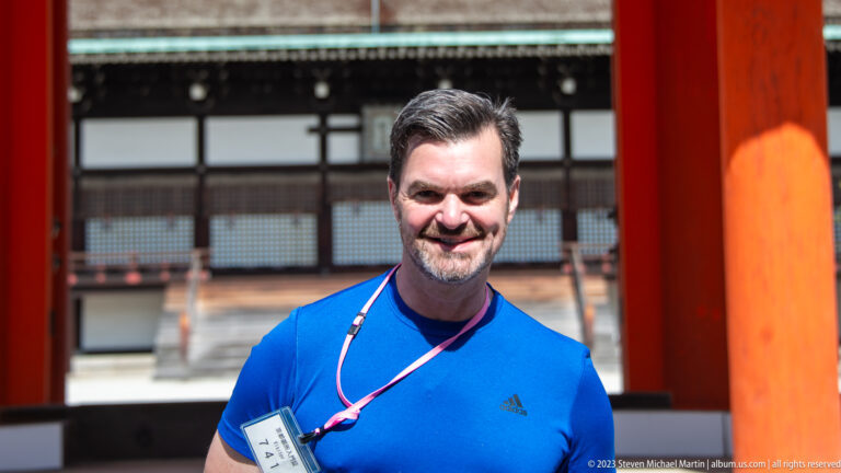 Close portrait of a man beside a vermilion post with a palace hall blurred behind.