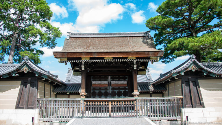 Frontal view of a wooden palace gate with a broad cypress-bark roof and flanking walls.