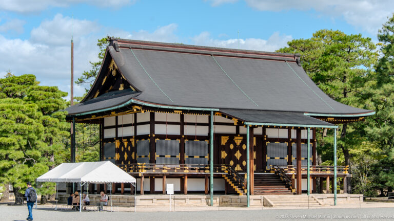 Raised palace pavilion with dark roof, patterned shutters, front steps, and pine trees.
