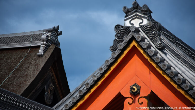 Close-up of a vermilion gable beneath ornate gray ridge tiles and overlapping palace roofs.
