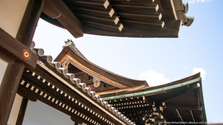 Upward view between palace eaves showing rafters, tiled roofs, and a curved gable.