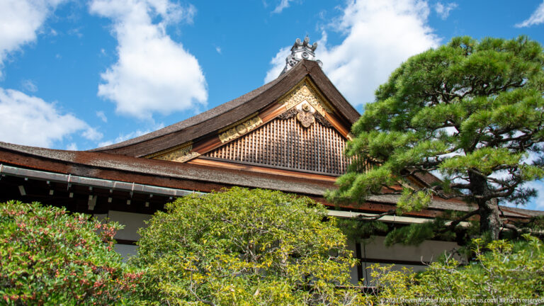Decorated palace gable with gold trim and louvered panel above garden foliage.