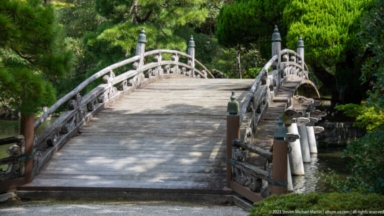 Arched wooden garden bridge with weathered rails surrounded by dense greenery.