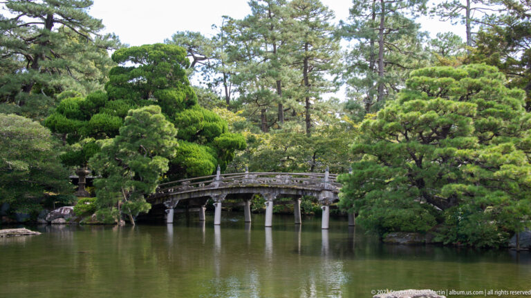 Arched wooden bridge crossing a pond among clipped pines and dense greenery.