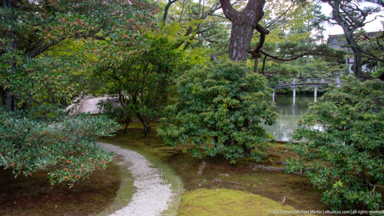 Winding gravel path through moss and shrubs with a pond and bridge visible beyond.