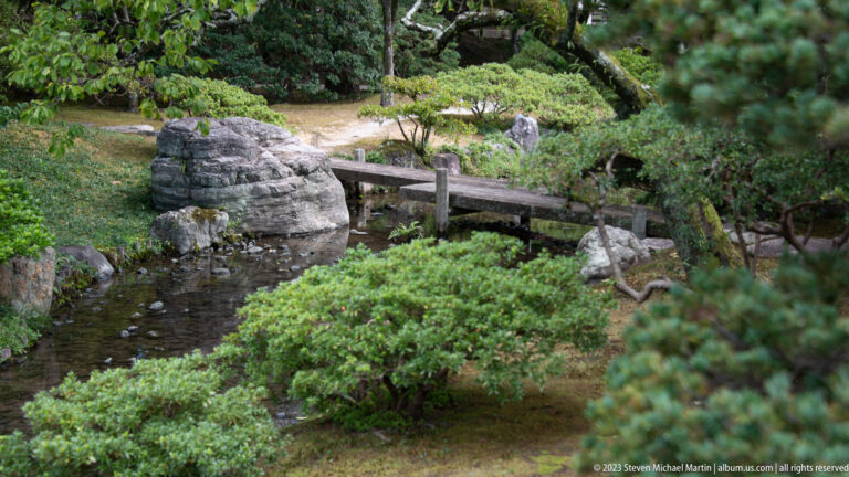 Narrow garden stream with rocks, clipped shrubs, and a simple wooden footbridge.