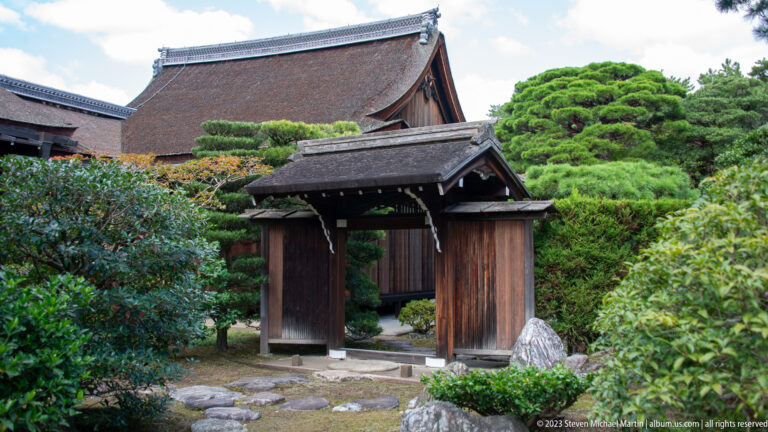 Small roofed timber gate amid shrubs with larger palace buildings behind.