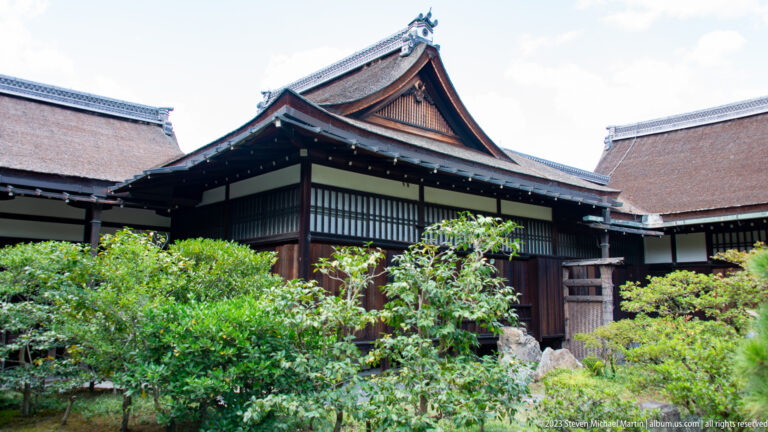 Gabled timber palace building with white panels partly screened by garden shrubs.