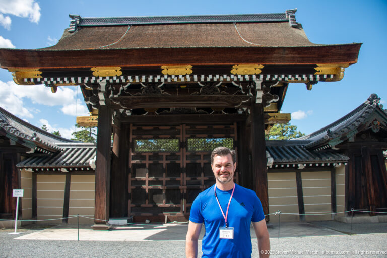 Man in a blue shirt standing before a large wooden gate at Kyoto Imperial Palace.