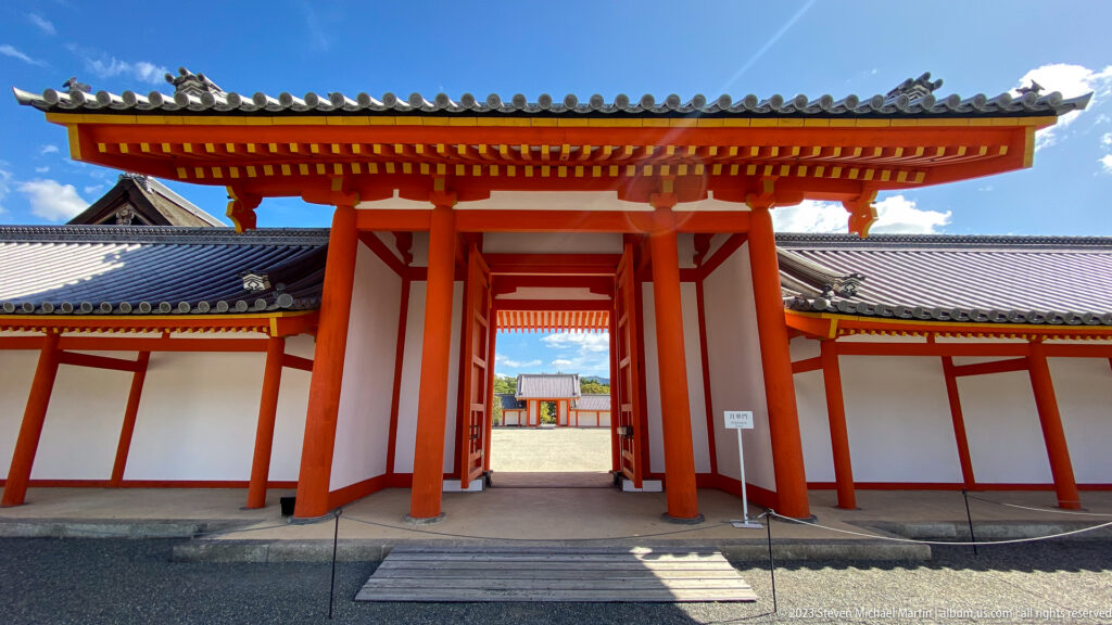 Open vermilion palace gateway framing a sunlit court and dark-roofed building.