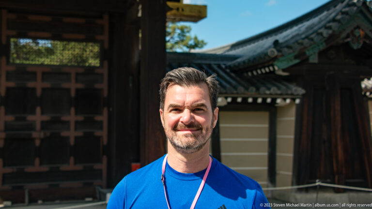Close portrait of a man in a blue shirt with a palace gate softly blurred behind him.
