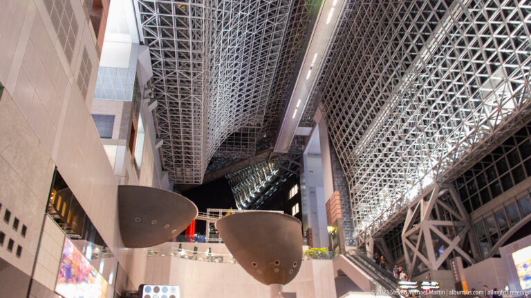 Three dark suspended pod-like forms above an interior terrace beneath Kyoto Station's steel roof.