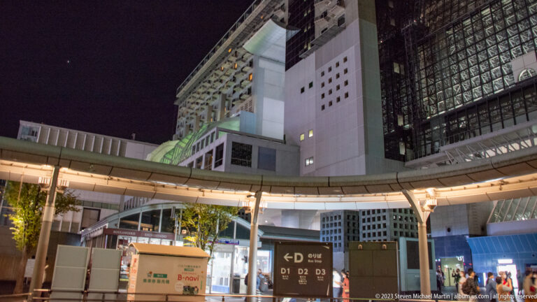 Curving elevated canopy in front of Kyoto Station's tall white walls and dark glazed facade at night.
