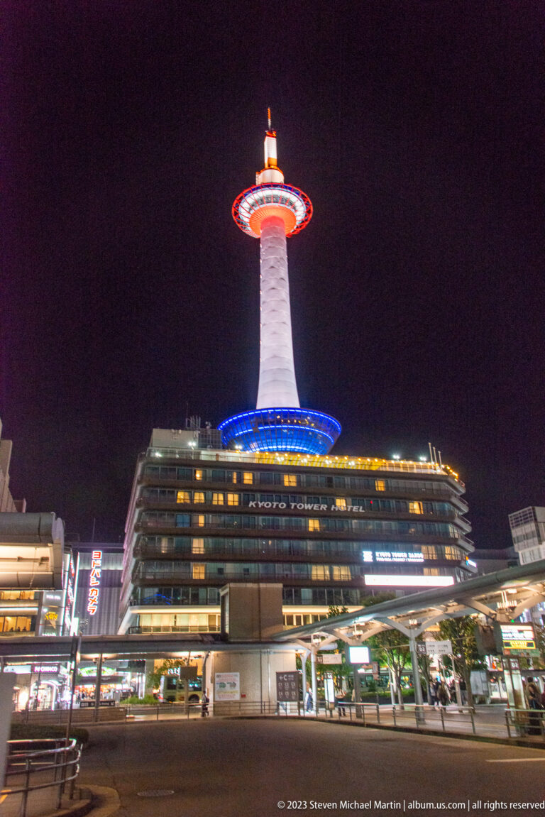 Wide night view of Kyoto Tower above its base building, with street traffic and storefronts below.
