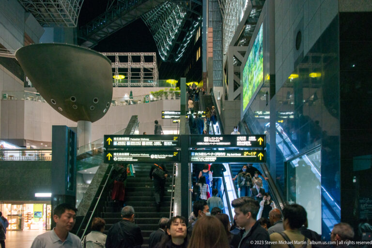 Travelers use escalators beneath suspended dark forms and steel roof framing inside Kyoto Station.
