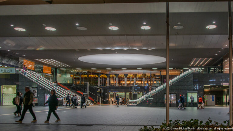 People cross an open Kyoto Station concourse behind glass, with escalators and illuminated shops in the background.