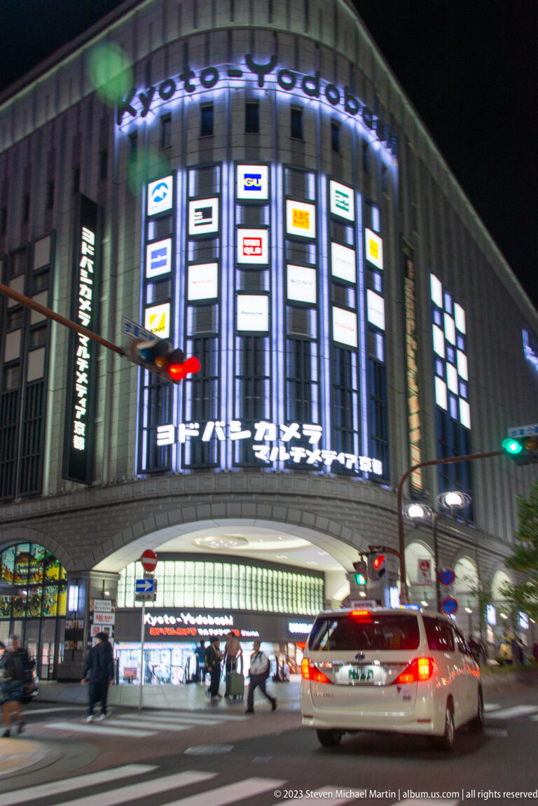 Night view of Yodobashi Kyoto's illuminated facade with pedestrians and a passing taxi.