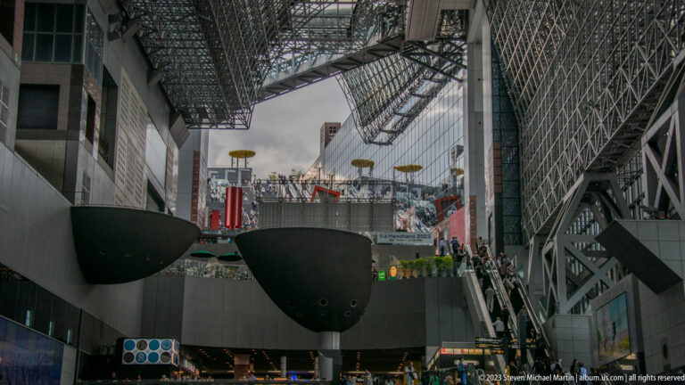High view across Kyoto Station's atrium with suspended forms, terraces, escalators, crowds, and steel roof.