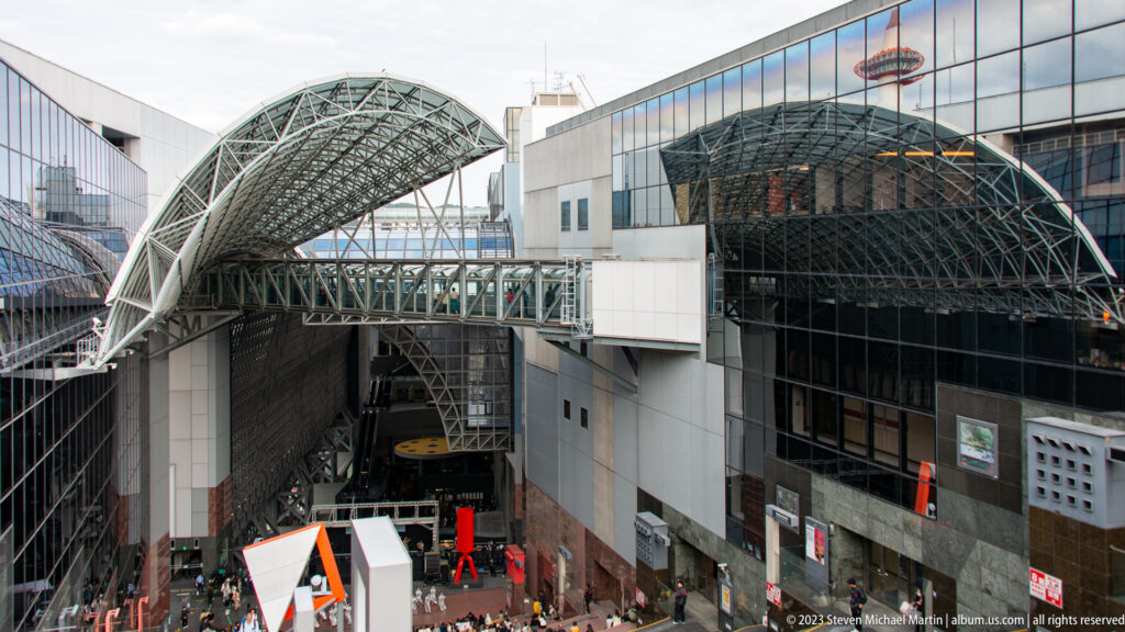 Elevated view of Kyoto Station's open plaza with stage, pedestrians, red sculpture, bridges, and curved roofs.