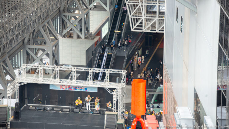 Elevated side view of a stage performance beside a large red sculpture within Kyoto Station's multi-level plaza.