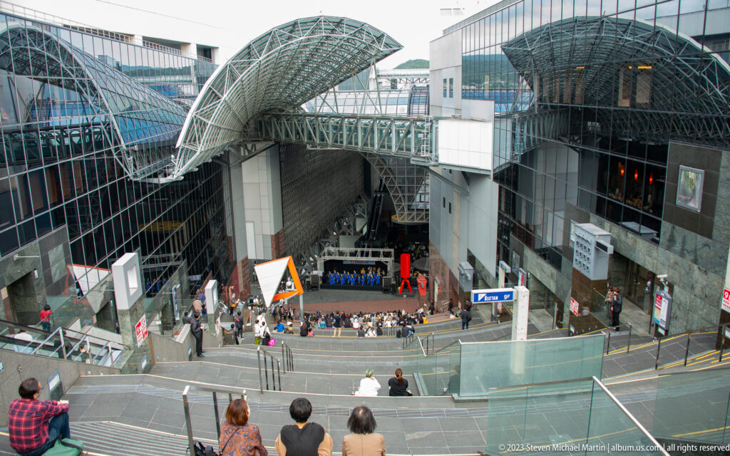 Elevated view of Kyoto Station's event plaza with a performance stage, pedestrian bridges, terraces, and curving roof structures.