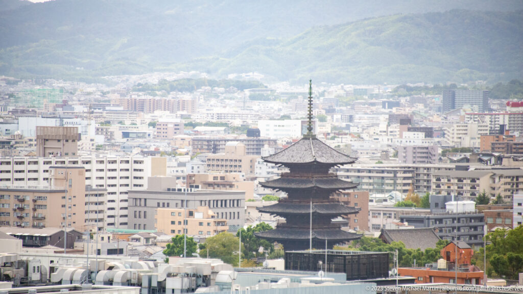 Telephoto view of Tō-ji's five-story pagoda rising above central Kyoto with mountains in the distance.