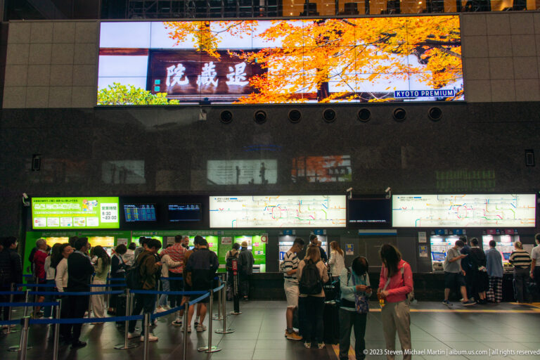 Passengers queue at Kyoto Station ticket counters beneath illuminated route displays and a large advertising screen.