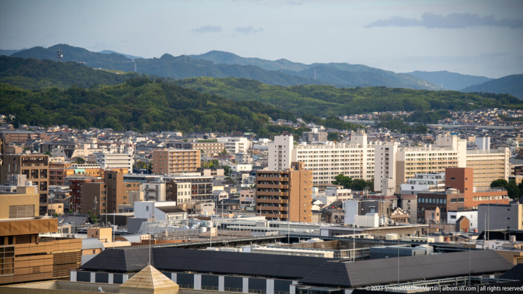 Elevated panorama of Kyoto with mid-rise buildings in the foreground and wooded hills beyond.