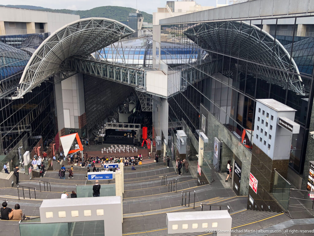 Elevated overview of Kyoto Station plaza with stage, bridges and roof.