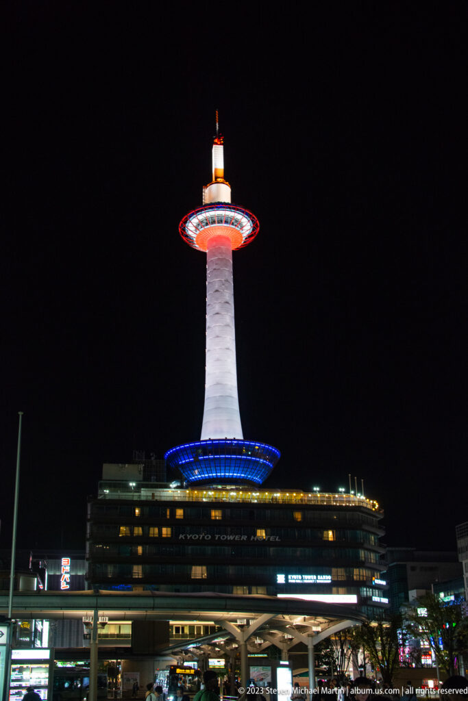 Kyoto Tower illuminated in white, blue, and red above station-side buildings at night.