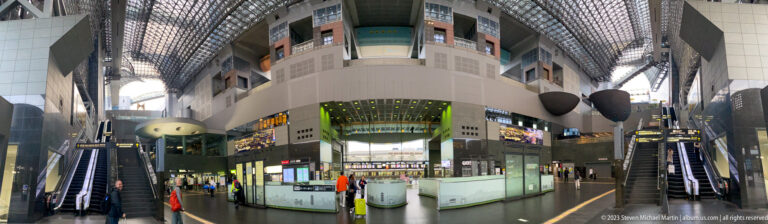 Wide view of Kyoto Station's central atrium with escalators, balconies, service counters, and a high steel-and-glass roof.