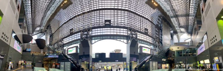 Vertical interior view of Kyoto Station with escalators, upper bridge, suspended forms, and a soaring glazed roof.
