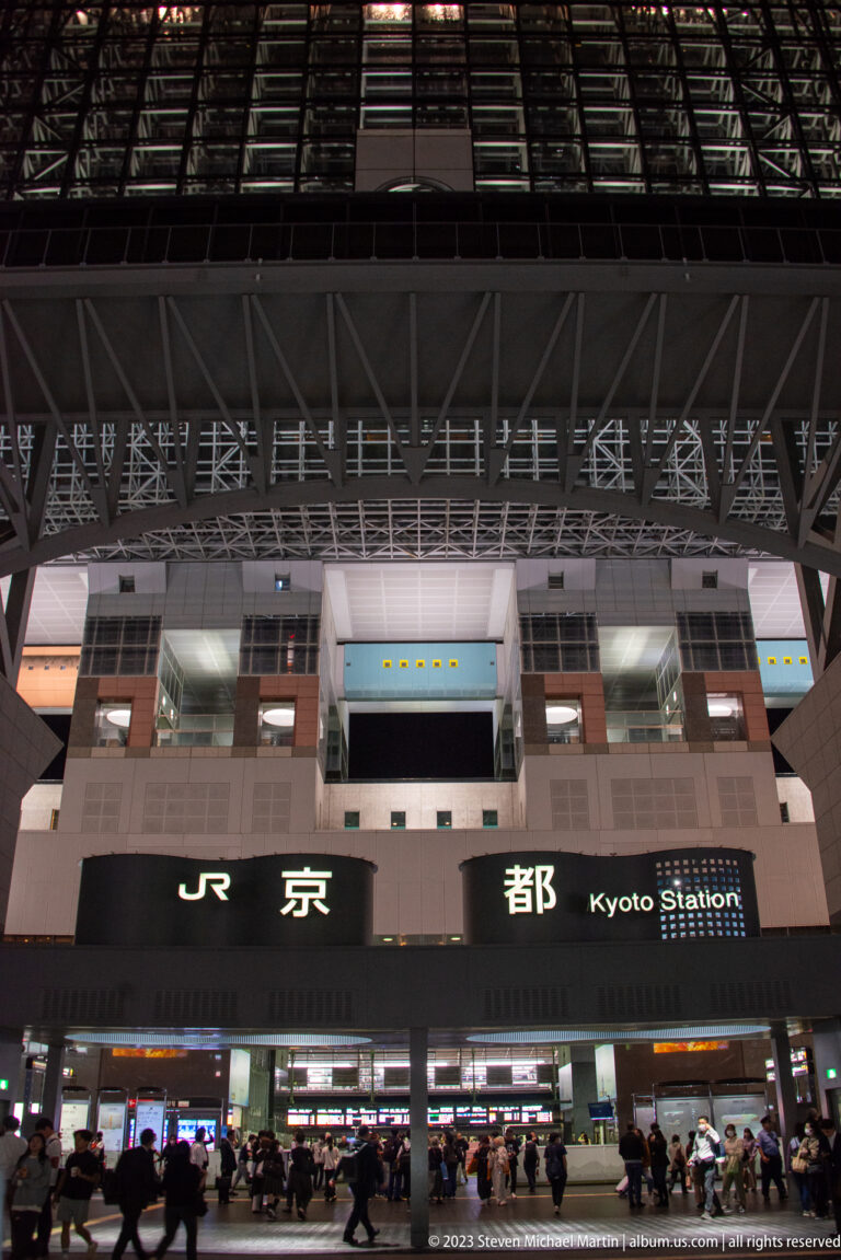 Night exterior of JR Kyoto Station with illuminated signage, entrance doors, and passengers in the forecourt.