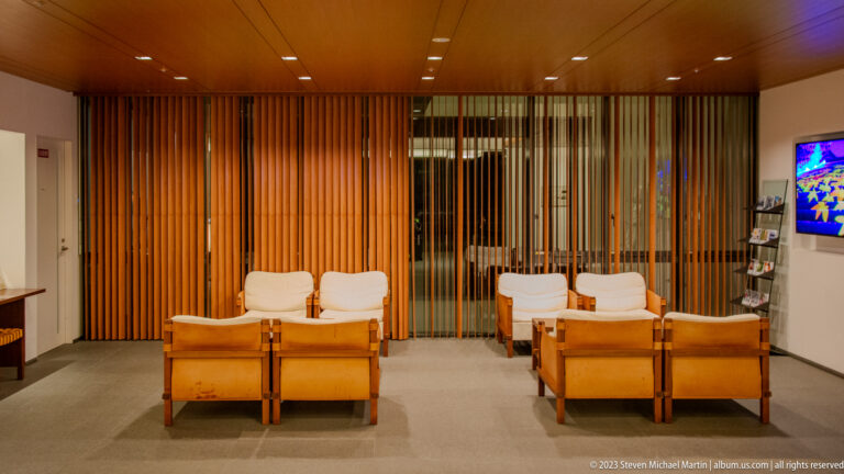 Modern hotel lounge with wooden armchairs, pale cushions, and tall vertical timber screens.