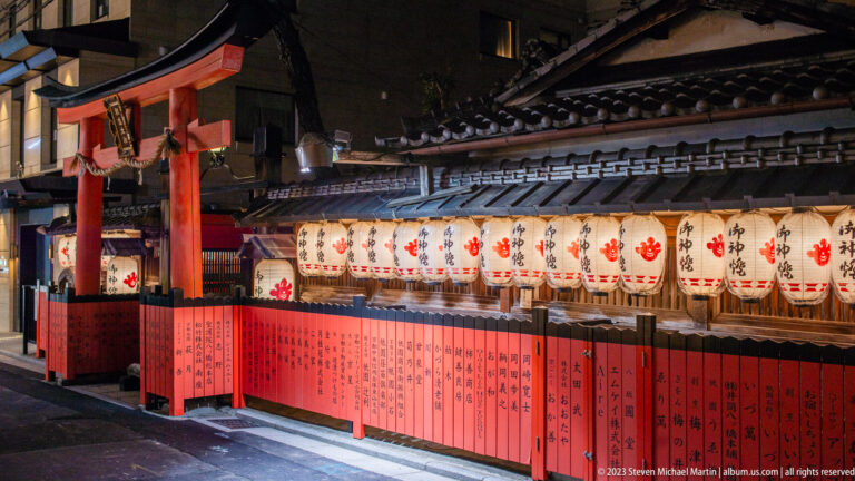 Shinto shrine enclosure at night with white lanterns, red inscribed panels, tiled roofs, and a small gate.