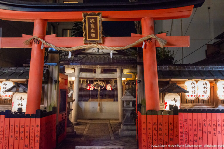 Red torii gate with sacred rope, paper streamers, lanterns, and a small shrine entrance beyond.
