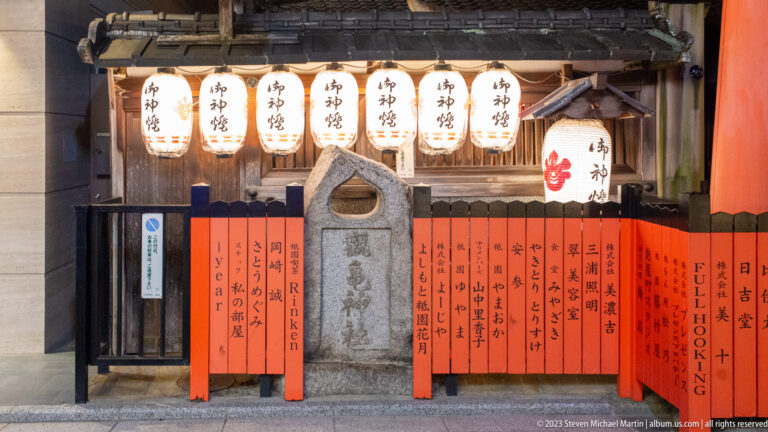 Carved stone shrine marker beneath white lanterns and behind red boards with vertical Japanese inscriptions.
