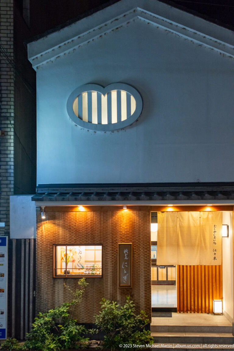 Contemporary Kyoto restaurant exterior with warm lighting, timber entrance, and oval clerestory window.