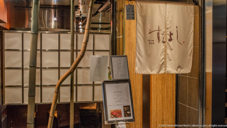 Restaurant entrance with noren curtain, bamboo pole, menu stands, and wood trim.