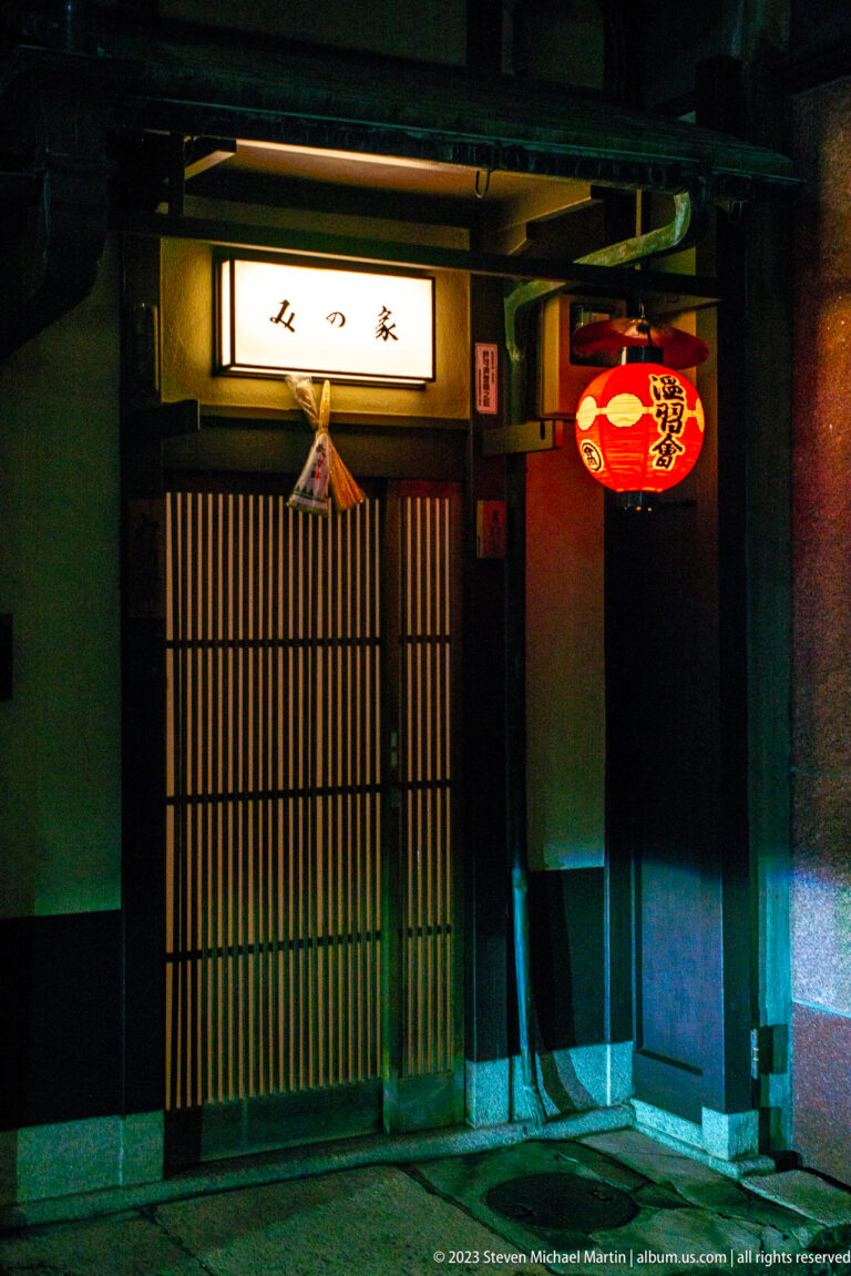 Dark restaurant entrance with wooden lattice doors, a red paper lantern, and a small illuminated sign.
