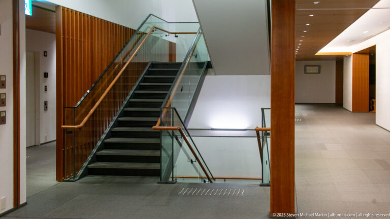 Interior staircase with dark steps, glass railings, wood handrails, and a bright adjoining corridor.