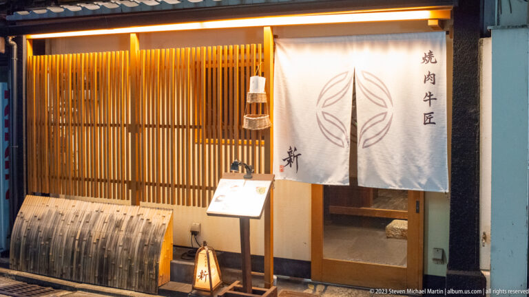 Yakiniku restaurant entrance with a white noren curtain, timber slats, and menu stand.