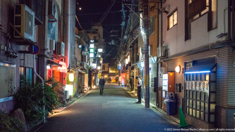 Narrow Kyoto alley at night with restaurant signs, lanterns, pedestrians, and a parked bicycle.