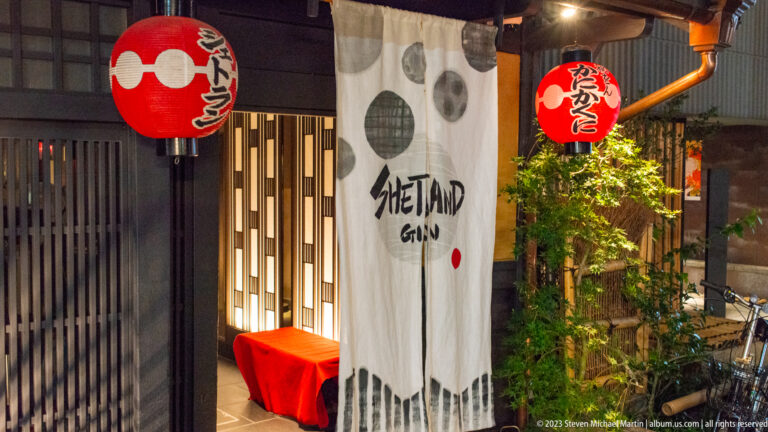 Gion restaurant entrance with white noren, red lantern, wood lattice, and greenery.